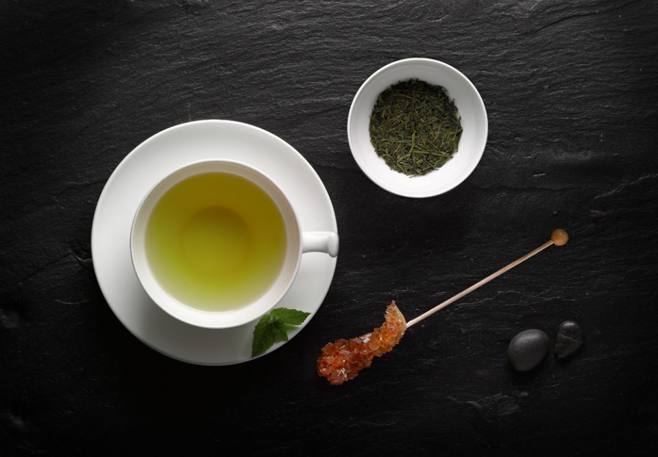 Tasse grüner Tee mit Kandis auf schwarzer Schieferplatte mit Natursteinen und Minze im Ambiente